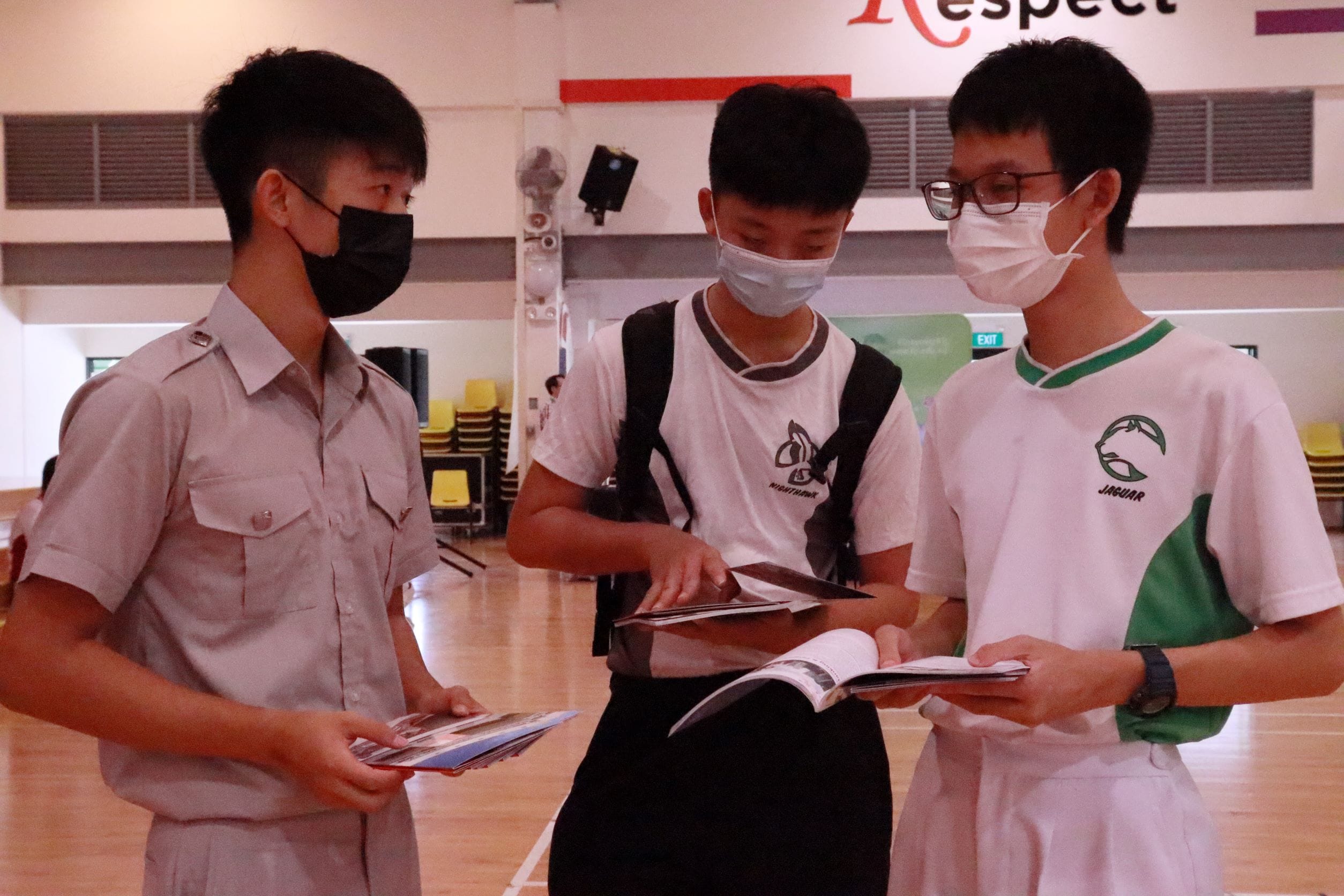 Three masked teenage boys stand in a school hall looking at printed booklets and pamphlets together.